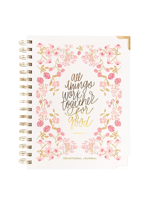 A spiral-bound devotional journal with a white cover decorated with pink watercolor flowers and gold foil lettering that reads 'all things work together for good'.
