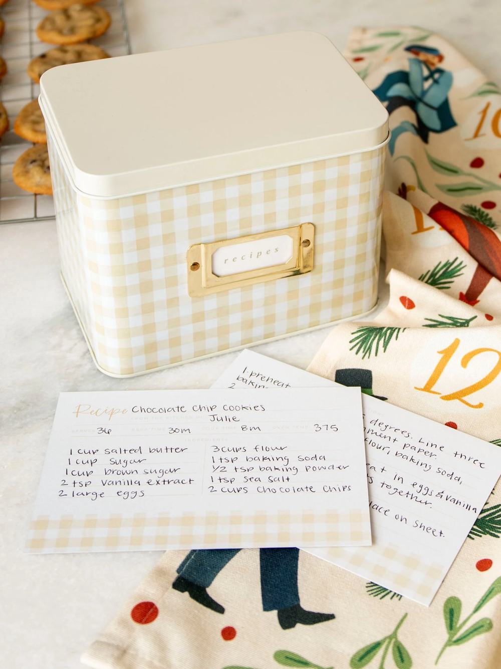 Beige gingham recipe tin box with metal label holder, shown with handwritten recipe cards on a kitchen countertop.