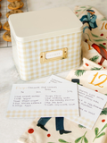 Beige gingham recipe tin box with metal label holder, shown with handwritten recipe cards on a kitchen countertop.