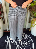 A person wearing black and white gingham wide-leg pants, standing on a dark doormat with potted plants on either side.