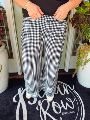 A person wearing black and white gingham wide-leg pants, standing on a dark doormat with potted plants on either side.