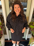 A smiling woman with long brown hair stands in front of a glass door wearing a black long-sleeve blouse with white polka dots and a black skirt.