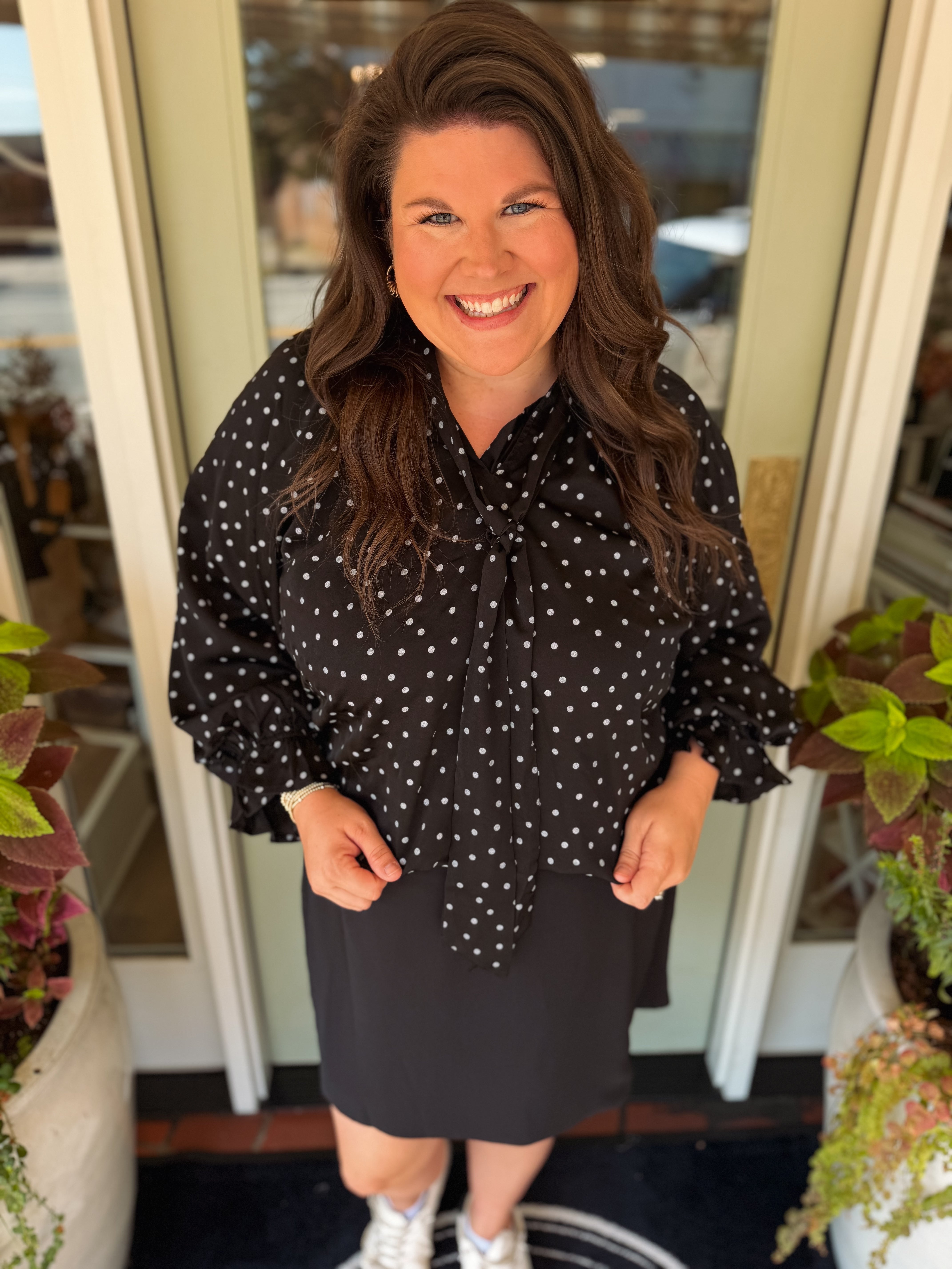 A smiling woman with long brown hair stands in front of a glass door wearing a black long-sleeve blouse with white polka dots and a black skirt.