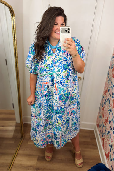 A woman takes a mirror selfie wearing a short-sleeve midi dress with a colorful abstract botanical pattern in blue, green, pink, and white. She is standing in a room with light wood floors, white walls, and a gold-framed mirror.