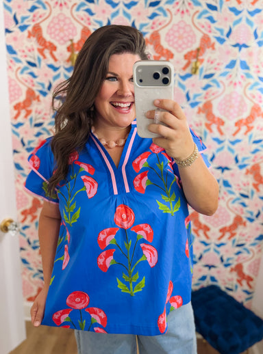 A woman takes a mirror selfie wearing a bright blue short-sleeve scrub top with a red and green floral pattern and pink trim. She is standing in front of a pink, blue, and orange patterned wall.