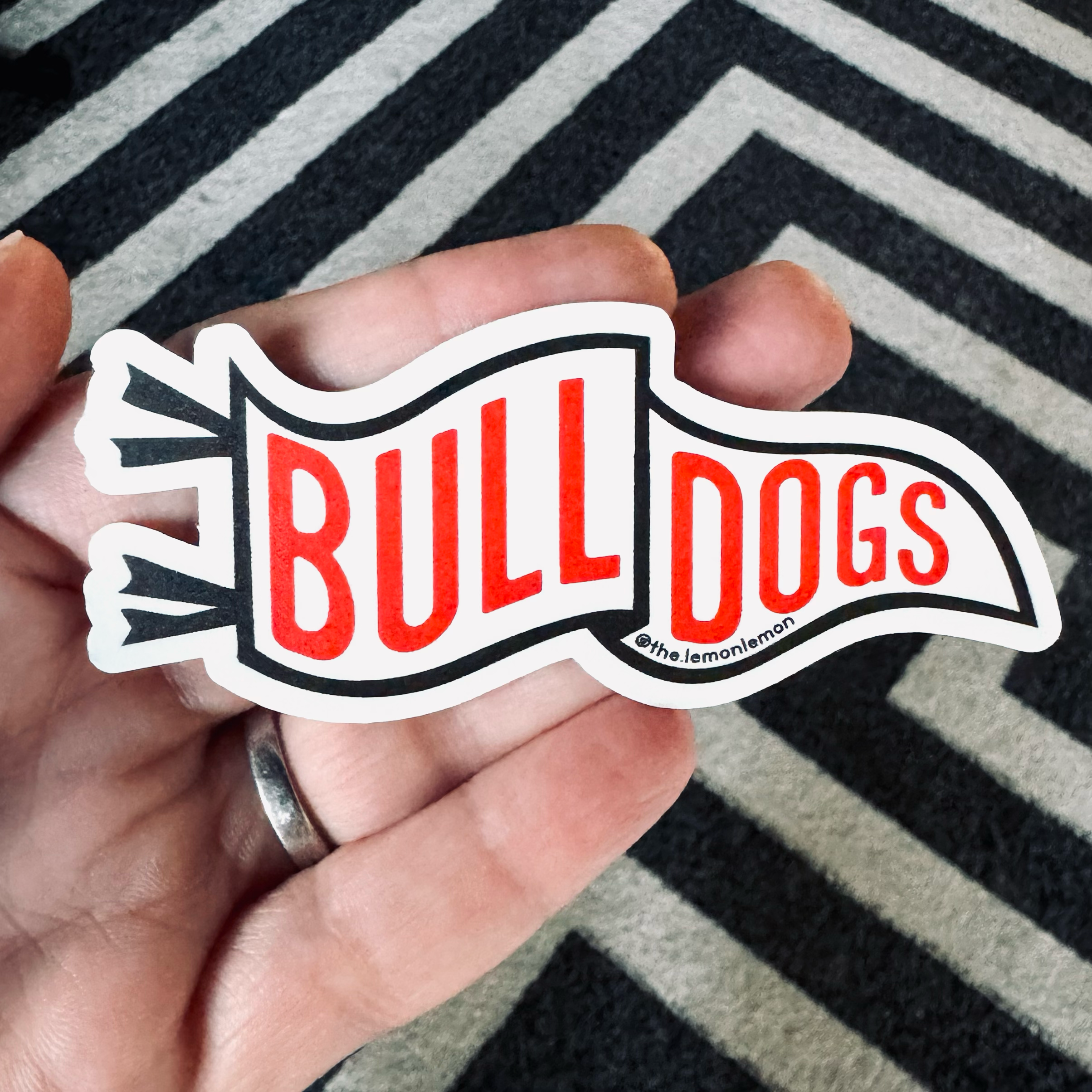 A hand holding a die-cut sticker that reads 'BULL DOGS' in red letters on a white banner with a black outline, set against a black and white striped rug.