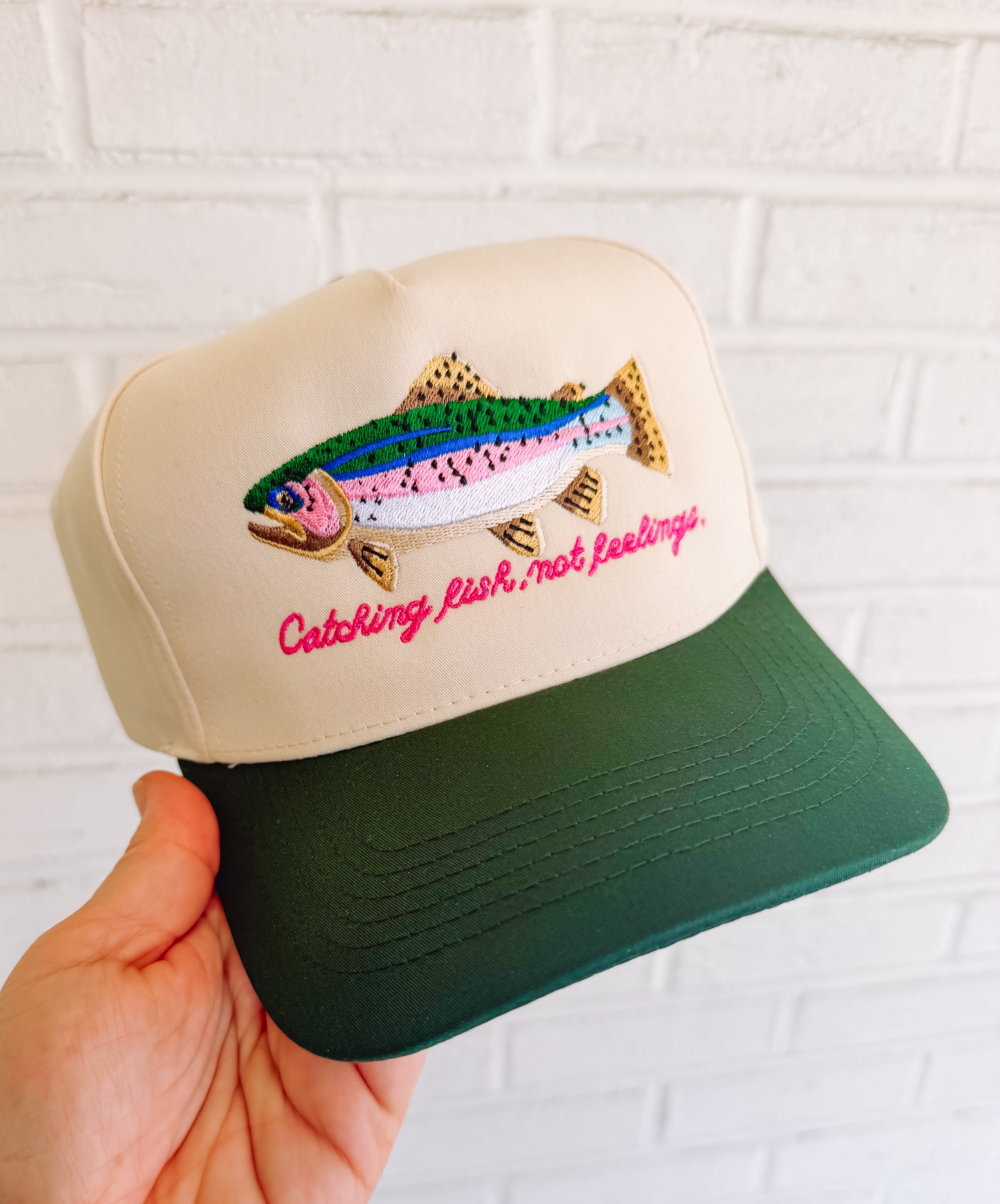 Trucker hat with embroidered rainbow trout and pink “Catching fish, not feelings” text with dark green bill.