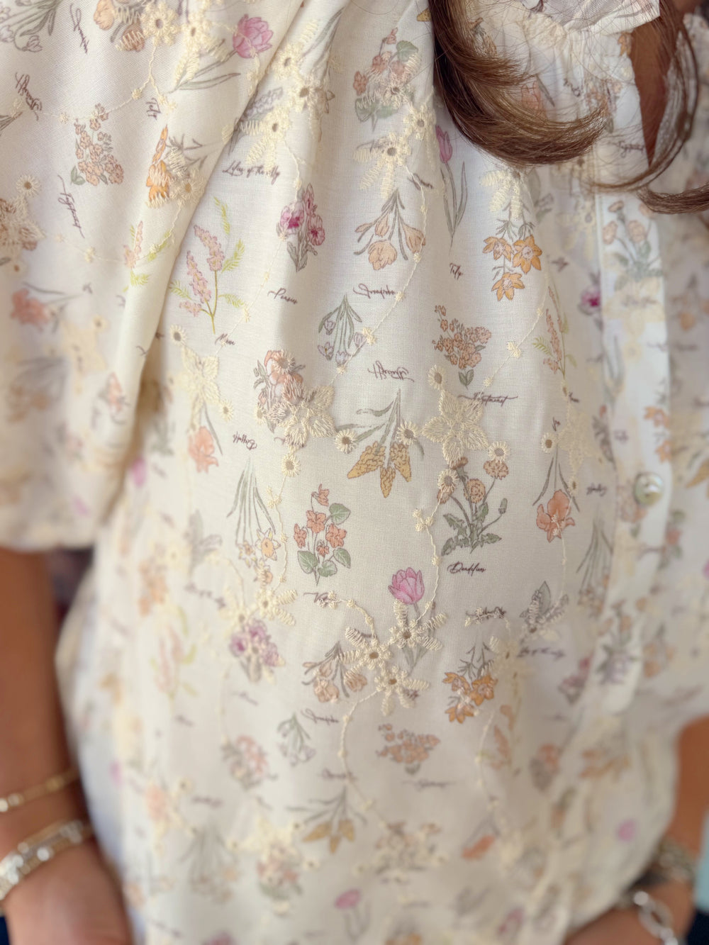 Woman wearing a cream floral print button-down blouse with short sleeves and ruffle neckline detail paired with light wash denim jeans.
