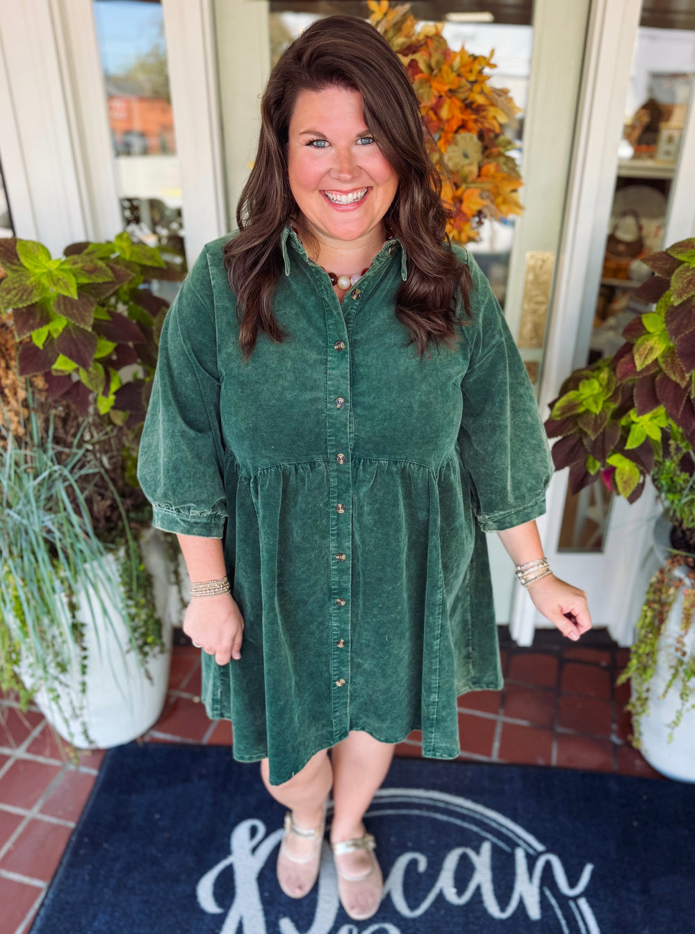 A woman wearing a dark green corduroy button-down dress stands smiling in front of a shop entrance decorated with potted plants and a fall wreath. The doormat features the text 'The Canopy'.
