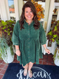 A woman wearing a dark green corduroy button-down dress stands smiling in front of a shop entrance decorated with potted plants and a fall wreath. The doormat features the text 'The Canopy'.