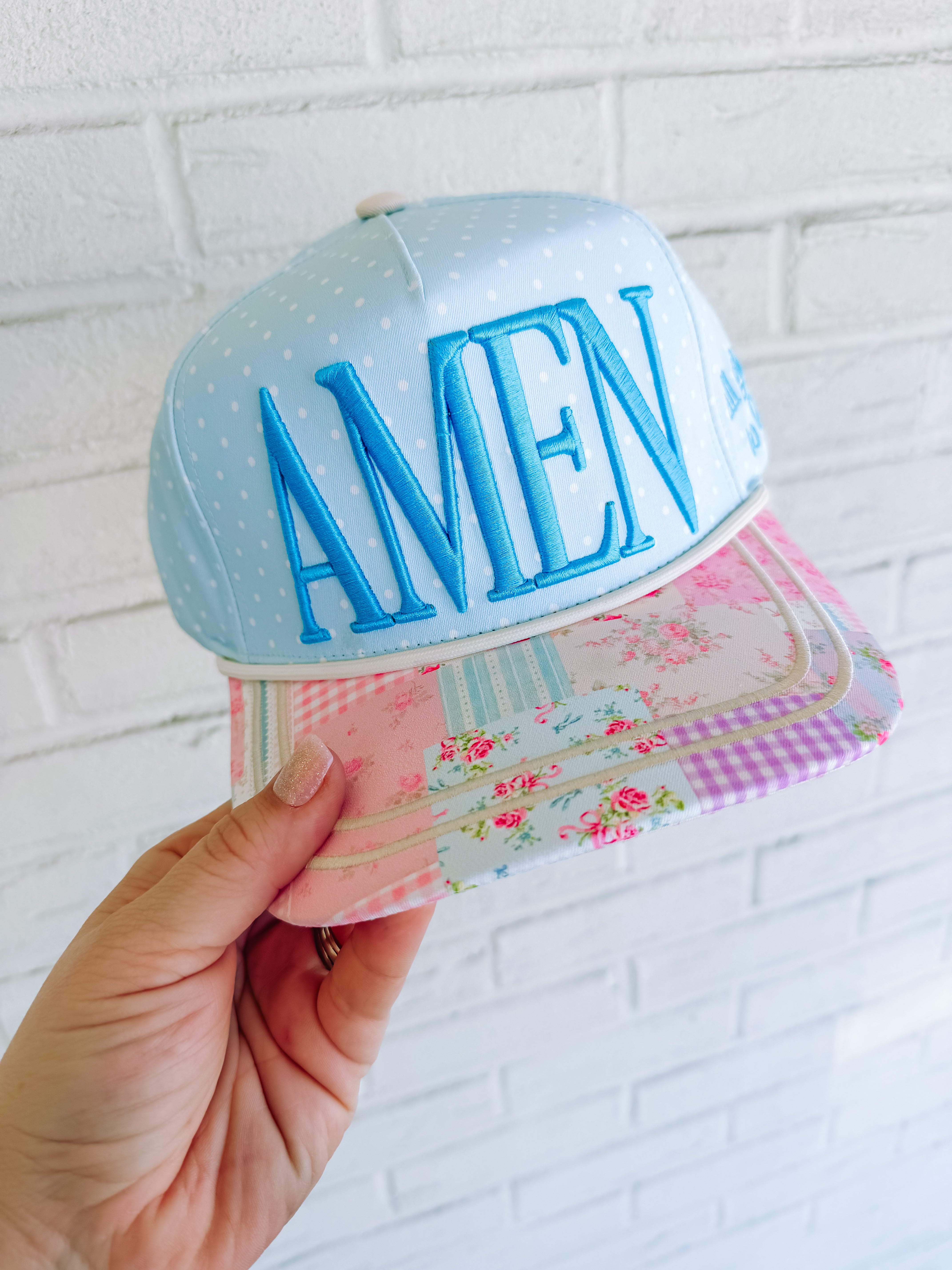 Colorful cap with 'AMEN' text held against a white brick wall.