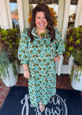A woman stands smiling in front of a doorway wearing a long-sleeve midi dress with a green, teal, and white botanical pattern. She is standing on a dark blue doormat with white script lettering, flanked by potted plants.