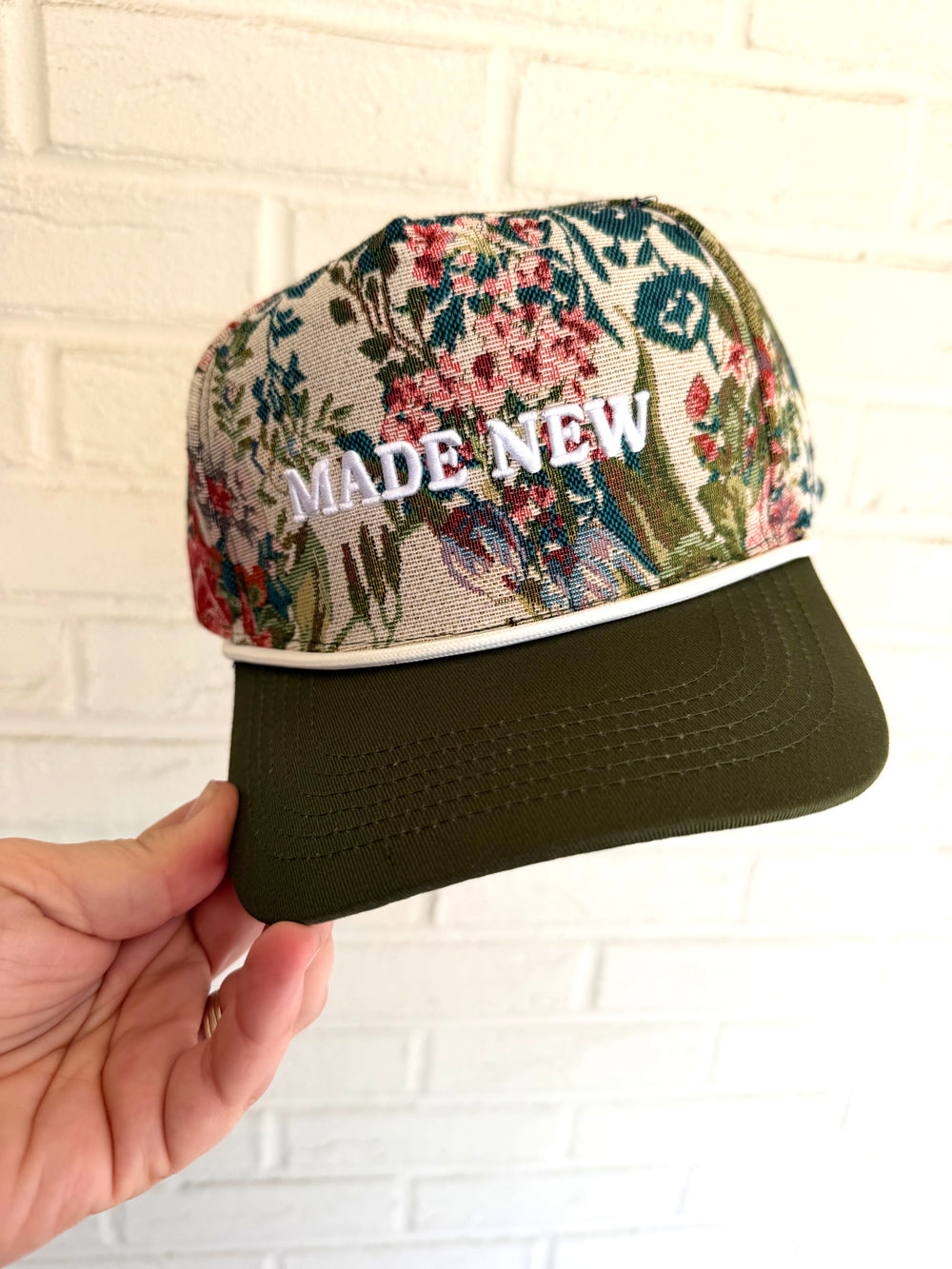Floral-patterned cap with 'MADE NEW' text held against a white brick wall.