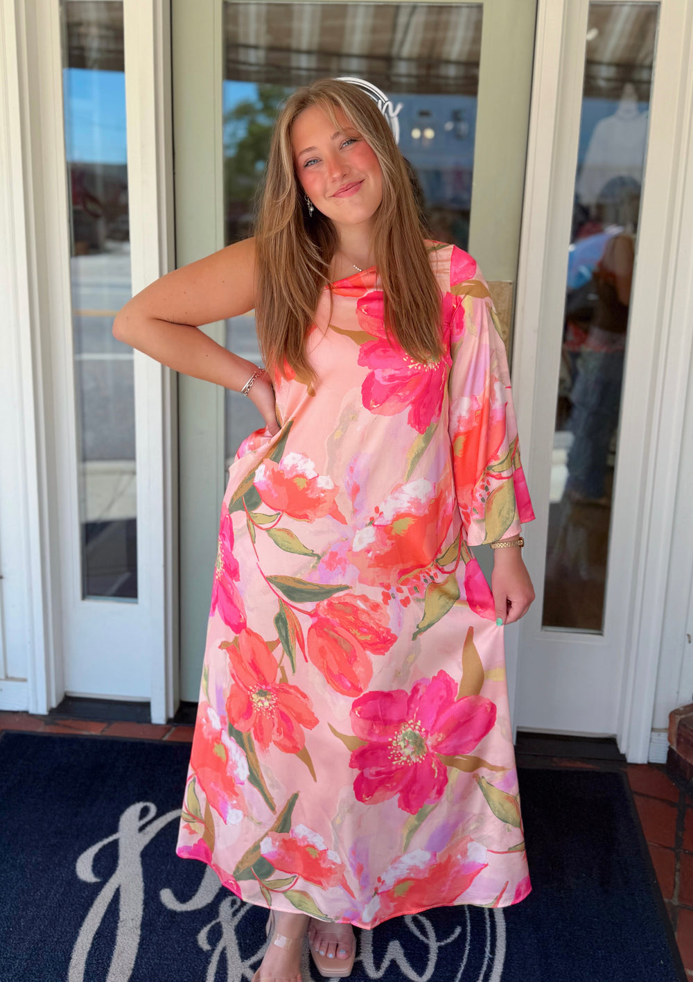 One-shoulder floral maxi dress featuring a blush pink background with oversized coral and hot pink watercolor flowers, crafted in a satin-finish fabric with a soft sheen.
