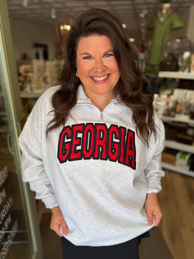 A woman smiling in a shop doorway wearing a light gray heathered quarter-zip sweatshirt with 'GEORGIA' printed across the chest in red and black arched lettering, paired with a black skirt.