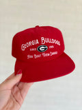 A red baseball cap with white embroidered text reading 'GEORGIA BULLDOGS', 'SINCE 1785', and 'How bout them dawgs' around a Georgia Bulldogs logo