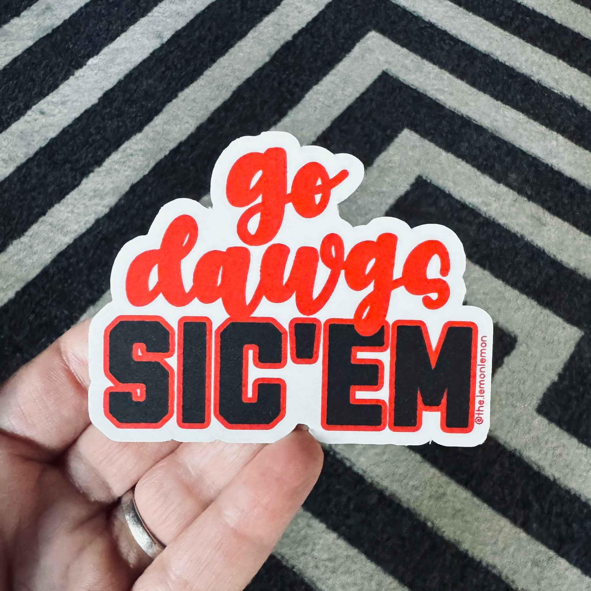 A hand holding a die-cut sticker that reads 'go daups SIC'EM' in red and black lettering with a white border, set against a black and gray chevron-patterned rug.