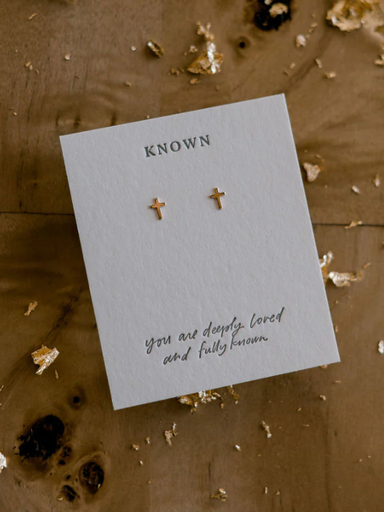 Gold cross earrings on a 'Known' branded card with a message on a textured surface.