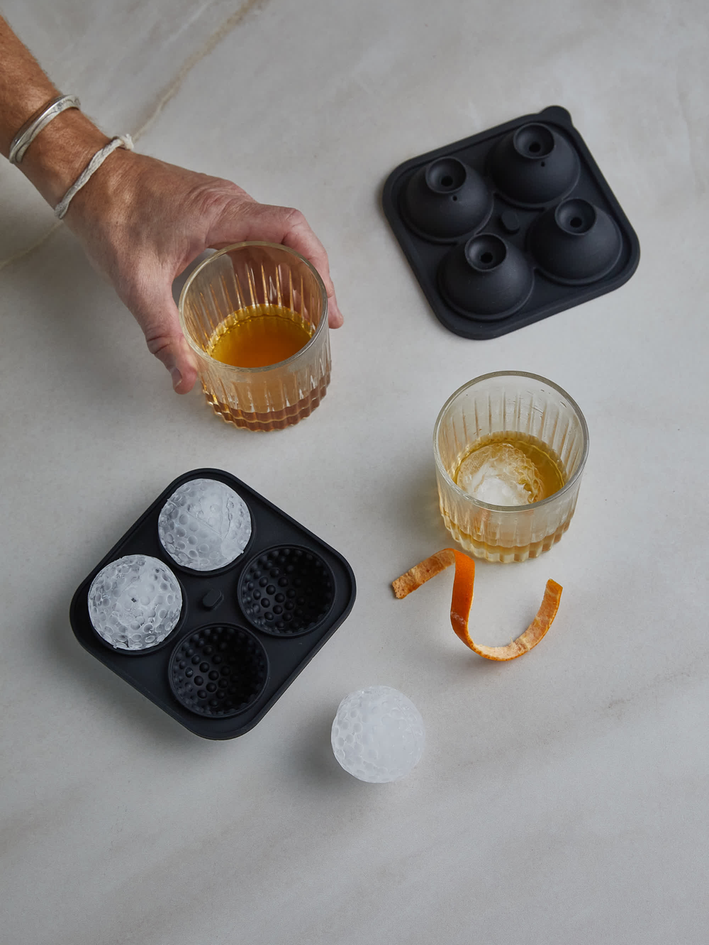 Black silicone ice cube tray with round and sphere molds on a light surface, with glasses of liquid and an orange peel.