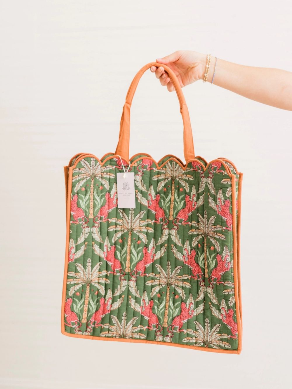 A hand holding a green patterned tote bag with orange handles and trim against a plain white background. The bag features a repeating botanical and bird print in green, pink, and beige.