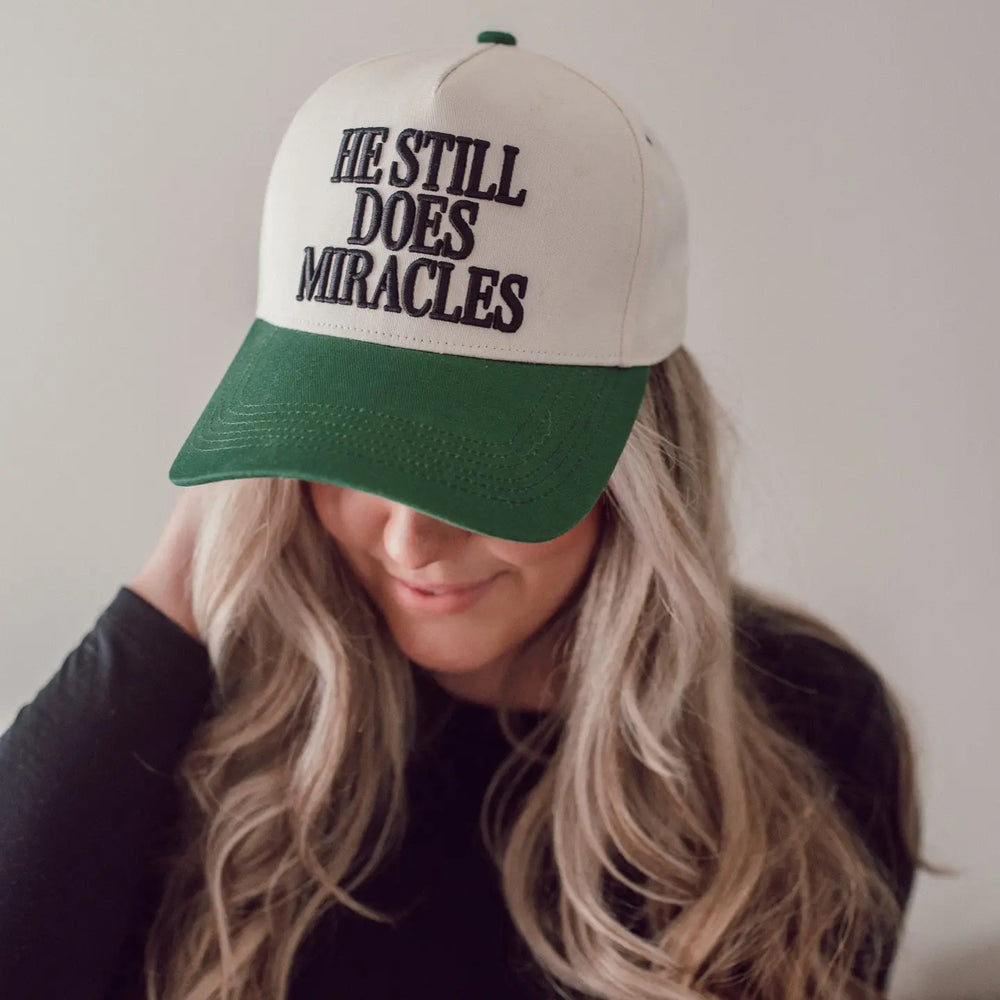 A woman wearing a beige and green baseball cap with the text 'HE STILL DOES Miracles' embroidered on the front panel.