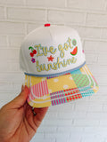 Colorful cap with 'I've got sunshine' text held against a white brick wall.