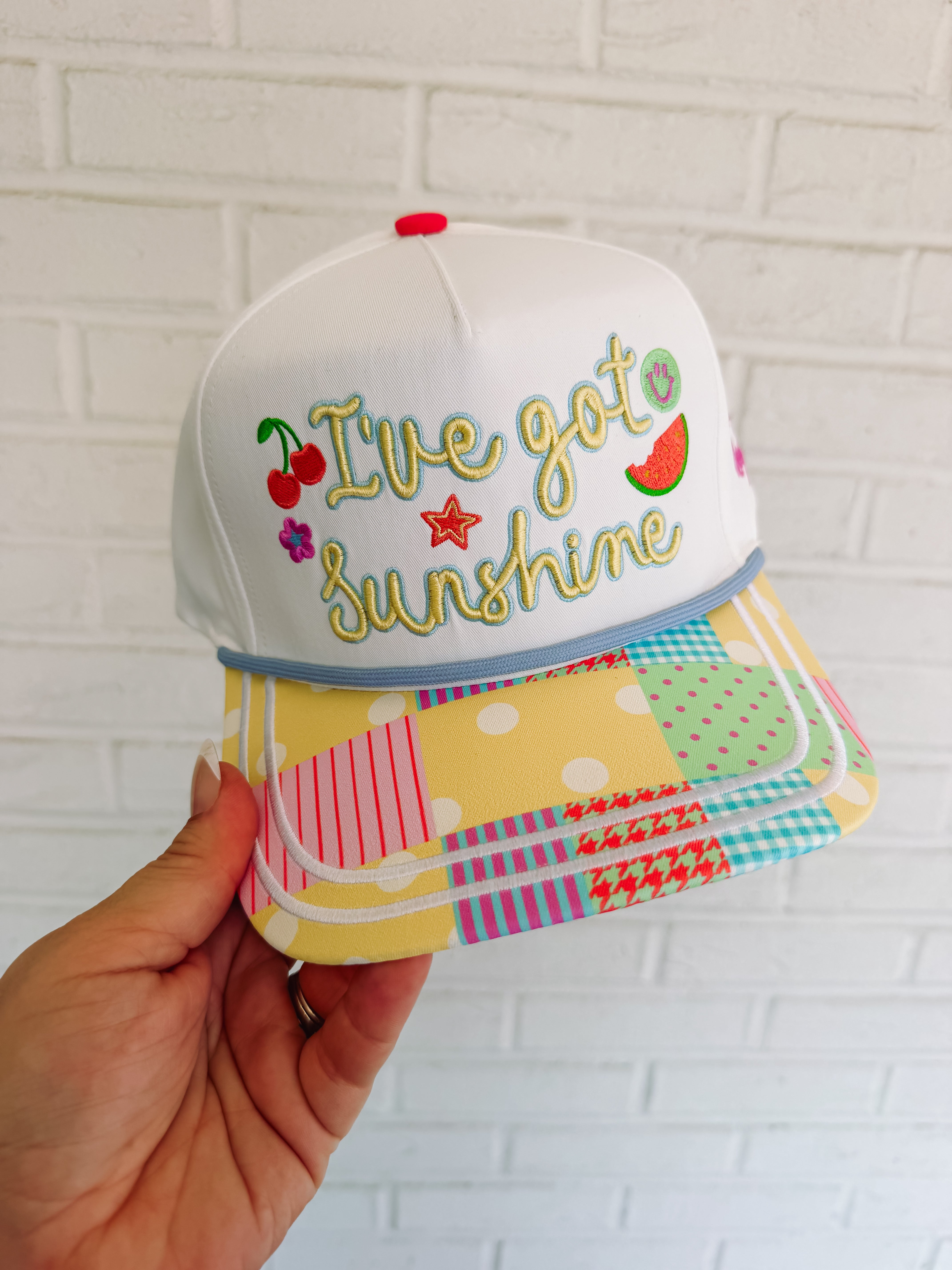 Colorful cap with 'I've got sunshine' text held against a white brick wall.