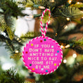A round pink cross-stitch Christmas ornament hanging from a decorated Christmas tree. The ornament features white embroidered text that reads 'IF YOU DON'T HAVE ANYTHING NICE TO SAY COME SIT BY ME' surrounded by small multicolored stitched dots, with a pink and white striped hanging loop.