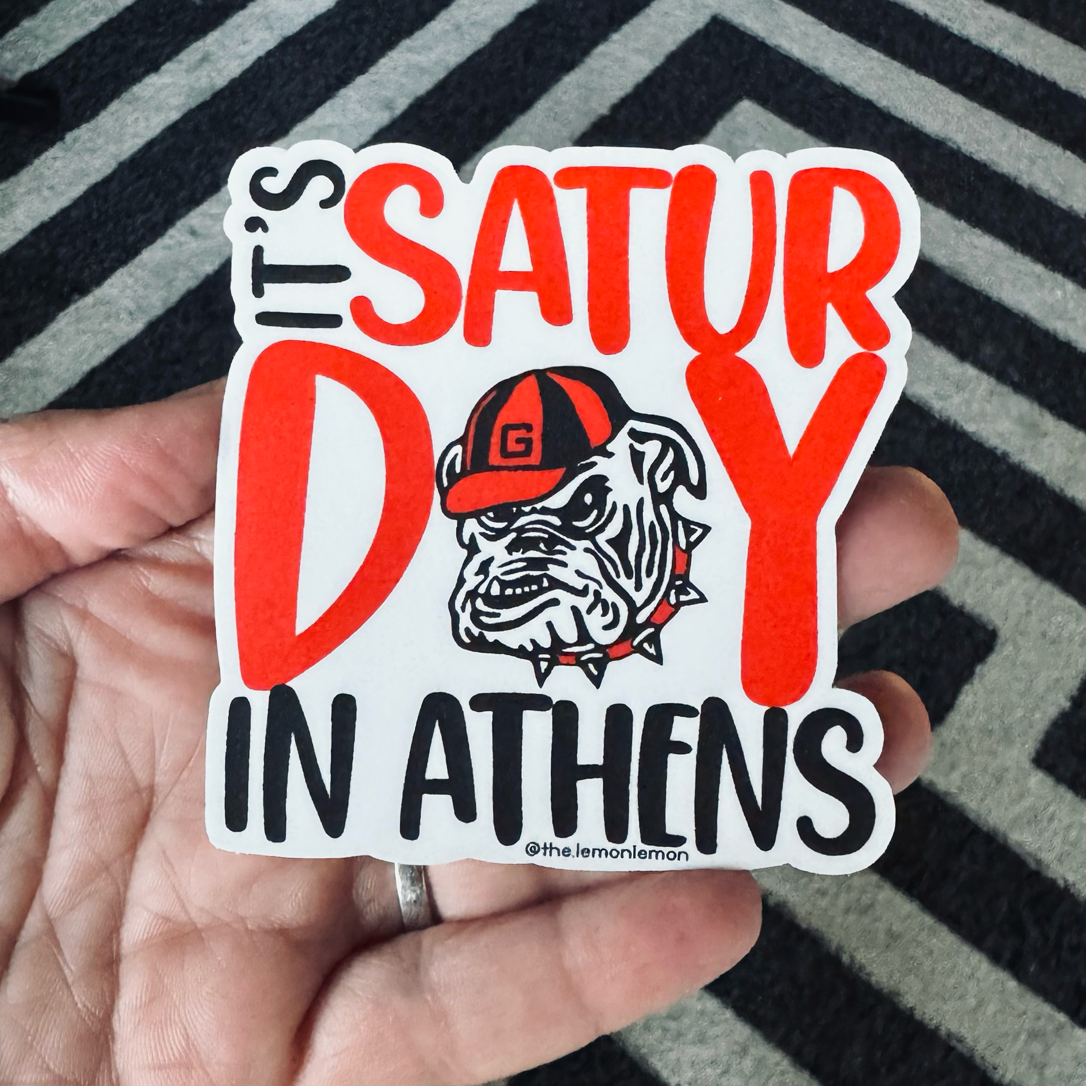 A hand holds a die-cut sticker that reads 'IT'S SATURDAY IN ATHENS' with a bulldog wearing a cap. The sticker is shown against a black and gray chevron-patterned rug.
