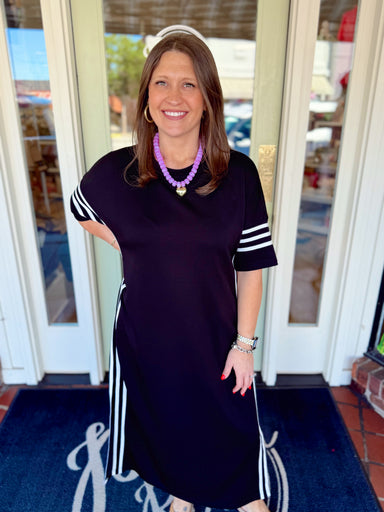 Woman wearing a black midi dress with white striped sleeve and side details paired with a purple beaded necklace and sandals.