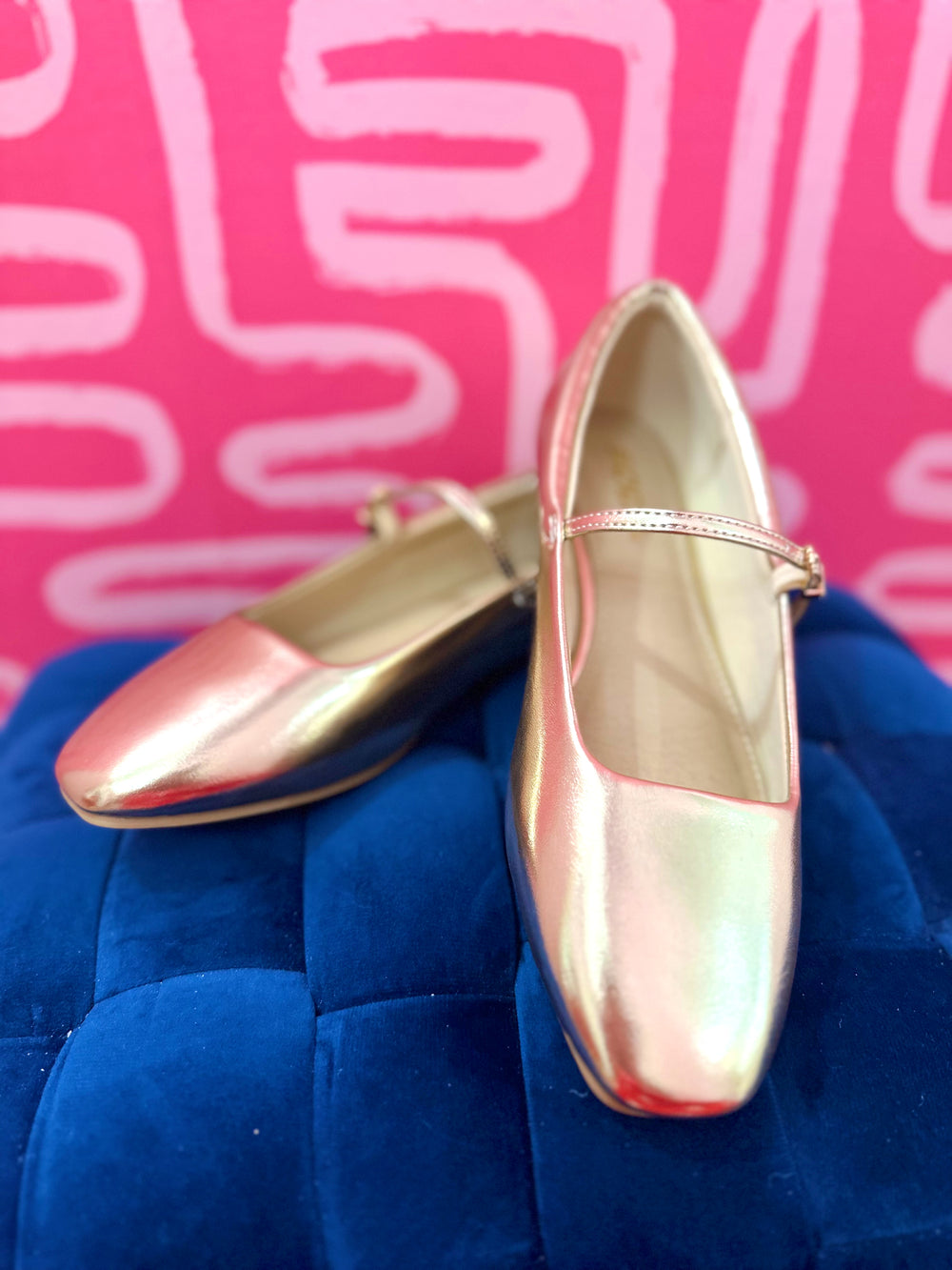 A pair of metallic rose gold Mary Jane flats resting on a blue velvet ottoman against a pink patterned background.