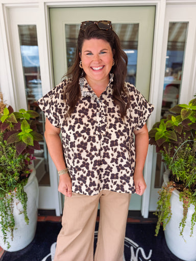 A smiling woman stands in front of a glass door wearing a short-sleeve leopard-print button-up shirt and tan pants, with potted plants on either side.