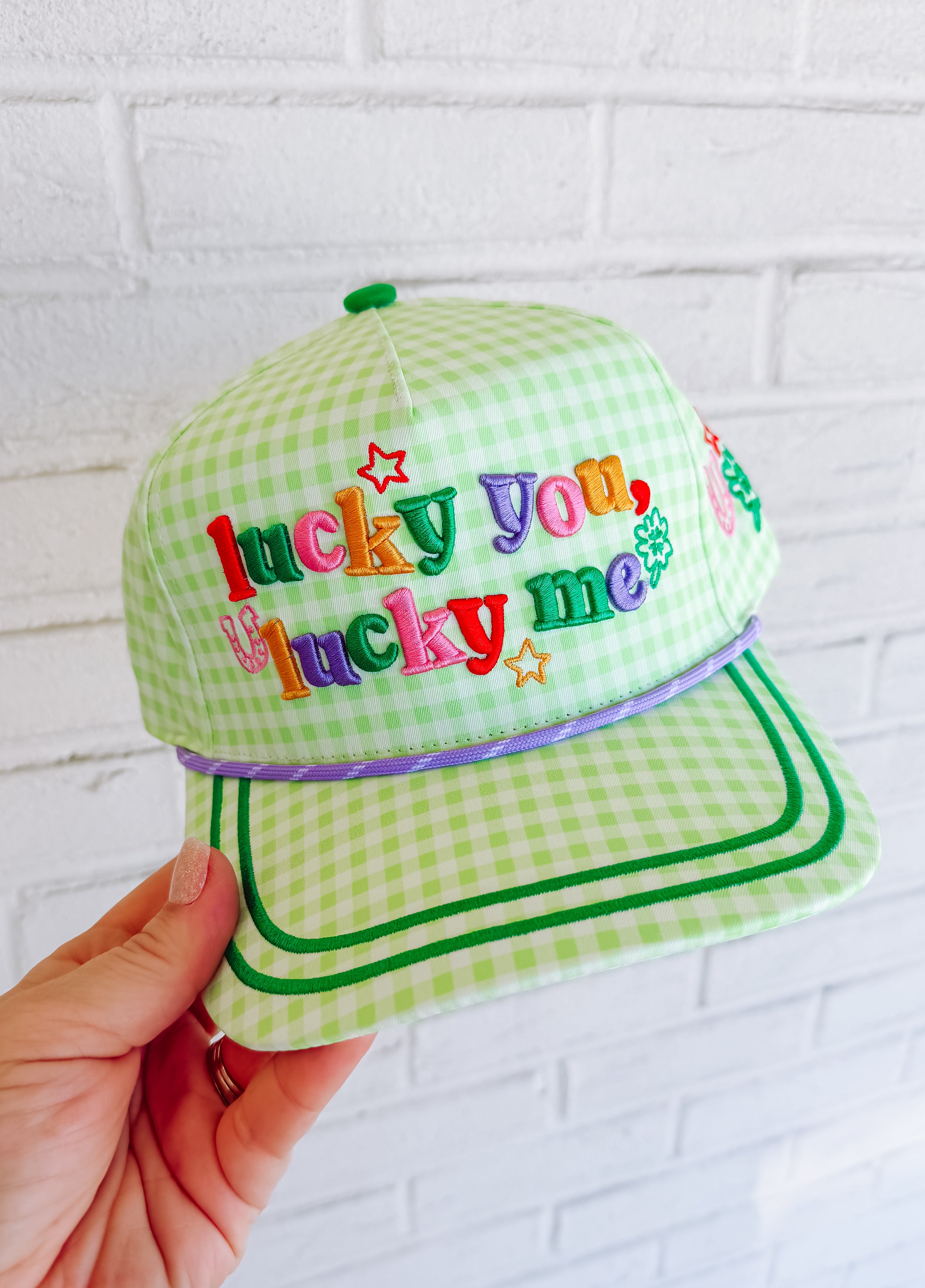 Green checkered cap with colorful text held against a white brick wall.