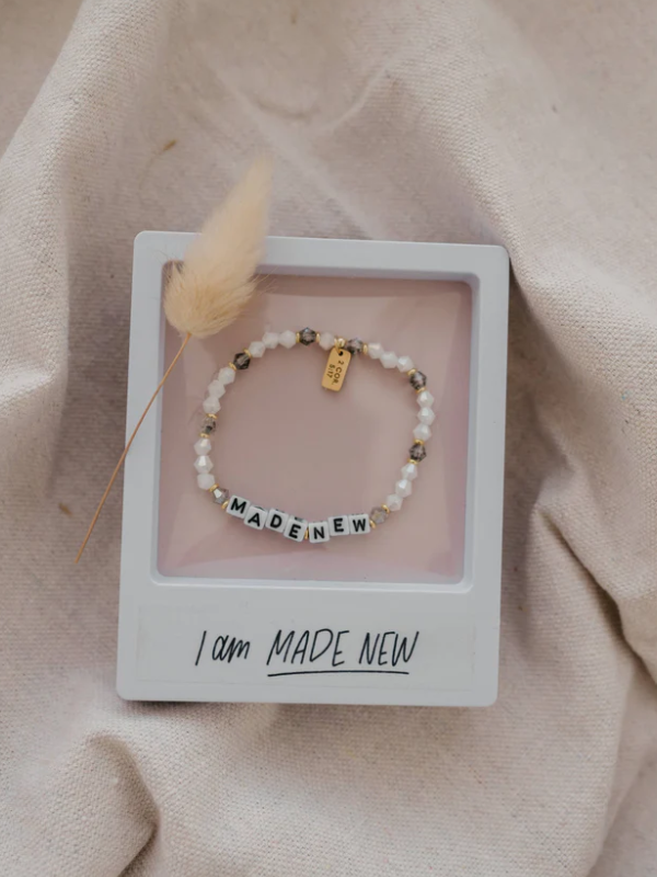 Bracelet with letter beads on a pink card with 'I am MADE NEW' text, on a beige fabric background.