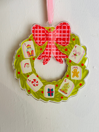 A festive Christmas wreath-shaped ornament hanging on a white wall. The ornament features a bright green scalloped border with red polka dots, a large red and white gingham bow at the top, and six numbered advent cards (1 through 6) attached around the wreath.