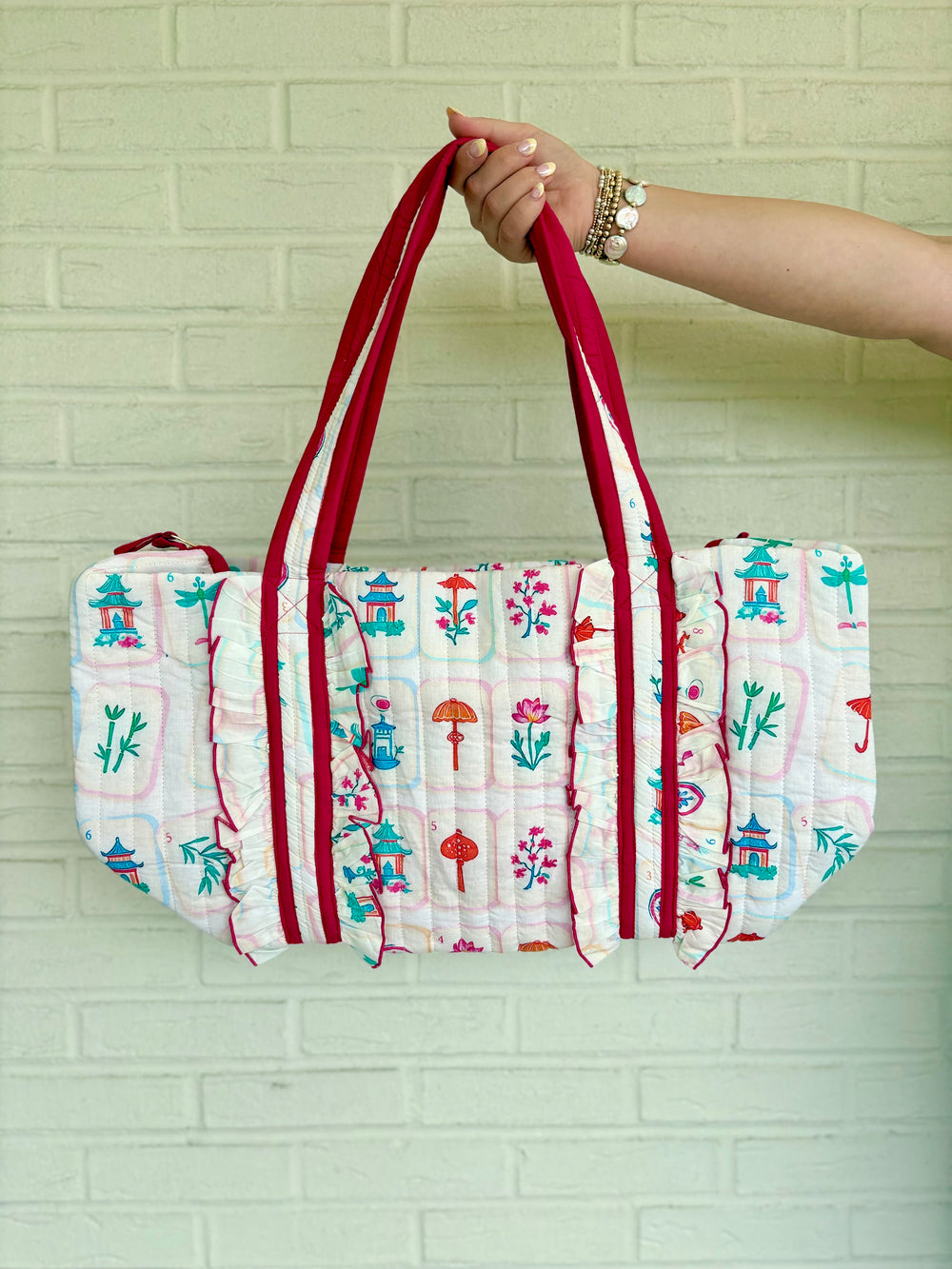 A quilted fabric tote bag with a white base, colorful printed motifs, and red straps, held by a person's hand against a light green brick wall.