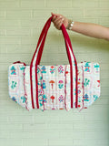 A quilted fabric tote bag with a white base, colorful printed motifs, and red straps, held by a person's hand against a light green brick wall.