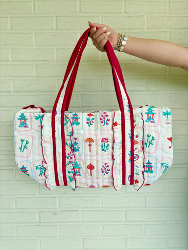 A quilted fabric tote bag with a white base, colorful printed motifs, and red straps, held by a person's hand against a light green brick wall.