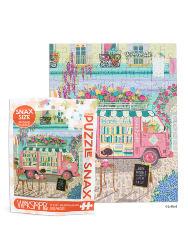 A colorful illustrated jigsaw puzzle and its retail box shown on a white background. The puzzle features a pink bookmobile filled with books, flowers, and outdoor seating, with the box labeled 'Puzzle Snax' and 'Wasserpill'.