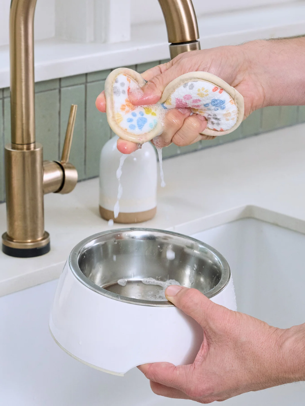 A person holds a white pet water bowl with a stainless steel interior under a brass faucet, while another person squeezes a floral-patterned pet lick mat above the bowl to wet it in a sink setting.