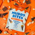 Muddy Bites peanut butter chocolate waffle cone snacks packaging on an orange background with scattered peanuts and chocolate pieces.