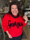 A woman smiling in a retail store wearing a bright red sweatshirt with 'Georgia' written across the front in black script, paired with a black skirt.