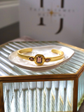 A gold cuff bracelet with a central rectangular pink gemstone and two smaller pink stones on either side, displayed on a white dish atop a ribbed glass table.