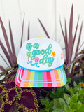 White trucker hat with colorful “It’s a Good Day” embroidery, rainbow striped brim, and beach-themed embroidered details including palm trees, waves, seashells, and starfish.
