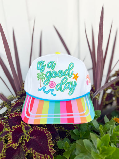 White trucker hat with colorful “It’s a Good Day” embroidery, rainbow striped brim, and beach-themed embroidered details including palm trees, waves, seashells, and starfish.
