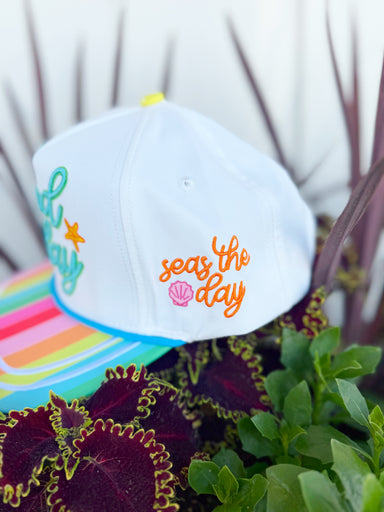 White trucker hat with colorful “It’s a Good Day” embroidery, rainbow striped brim, and beach-themed embroidered details including palm trees, waves, seashells, and starfish.
