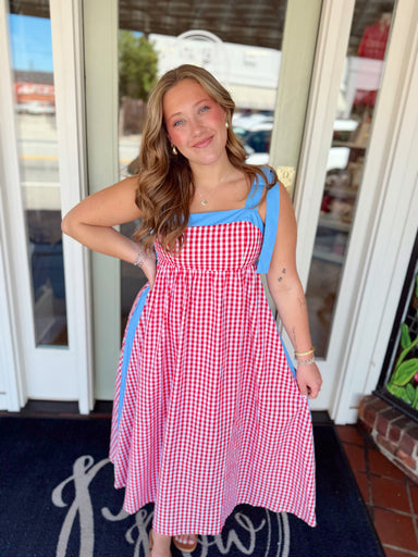 Woman wearing a red and white gingham midi dress with blue tie shoulder straps and blue trim detail paired with sandals and delicate jewelry.