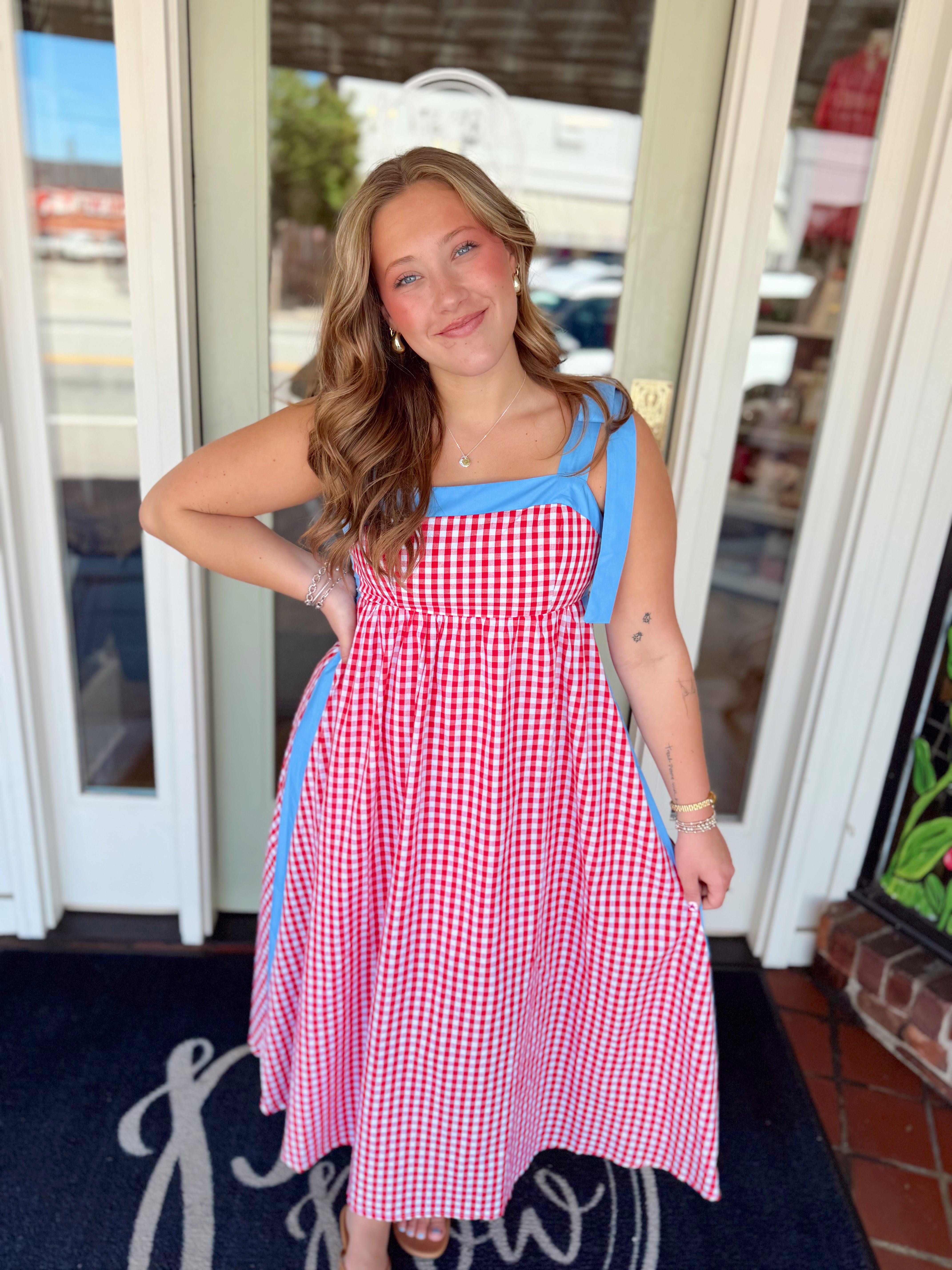 Woman wearing a red and white gingham midi dress with blue tie shoulder straps and blue trim detail paired with sandals and delicate jewelry.