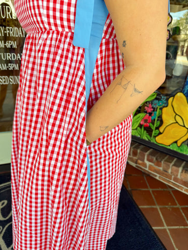 Woman wearing a red and white gingham midi dress with blue tie shoulder straps and blue trim detail paired with sandals and delicate jewelry.