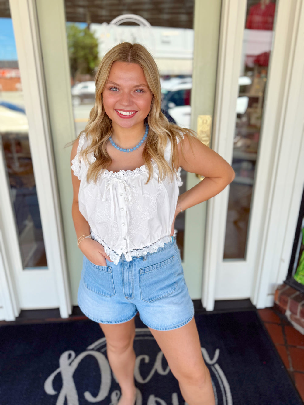 Woman wearing a white embroidered lace trim tank top with front tie detail paired with light wash denim shorts and a blue beaded necklace.